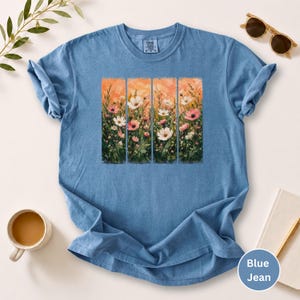 May include: Blue jean-colored t-shirt featuring a floral design. The design is a vertical panel of flowers in shades of pink, white, and green against an orange background. The shirt has short sleeves and a crew neck.