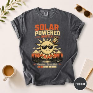 May include: Dark gray t-shirt with the text "SOLAR POWERED (MOSTLY)" above a cartoon sun wearing sunglasses. The sun is charging a phone with a lightning bolt. Below the sun is the text "Charging... Please Wait."