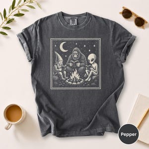 May include: Dark gray t-shirt featuring a square graphic of a gnome, Bigfoot, and an alien reading around a campfire under a crescent moon and stars. The shirt is a Pepper color.