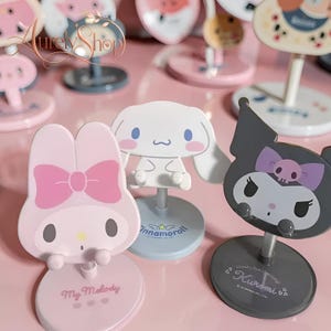 Pode incluir: Uma coleção de suportes para telemóveis com temas de personagens. Os suportes apresentam designs de My Melody, Cinnamoroll e Kuromi. Cada suporte tem uma base redonda e uma haste de metal. O suporte My Melody é rosa com um laço, o Cinnamoroll é branco e o Kuromi é cinzento.