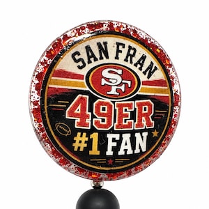 May include: A circular badge featuring a red glitter border and the text SAN FRAN 49ER #1 FAN with a football logo. Below the badge is a black matte bead and a brown football-shaped bead, forming a decorative accessory for a badge reel or pen.