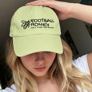 May include: A light yellow baseball cap featuring a black graphic of a bee and the text FOOTBALL HONEY 100% GAME DAY READY printed on the front. The cap has a structured crown, a curved brim, and stitched eyelets for ventilation.