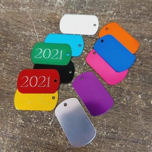 May include: A collection of rectangular metal dog tags in various colours including white, orange, blue, pink, purple, black, yellow, green, and silver. Two of the tags, one green and one red, are engraved with the year 2021 in a serif font.