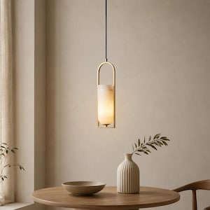 Może przedstawiać: Cylindryczna lampa wisząca z kloszem z białego kamienia emitującym ciepłe światło, zamknięta w ramie ze szczotkowanego mosiądzu. Oprawa wisi na cienkim czarnym przewodzie nad okrągłym drewnianym stołem z żebrowanym ceramicznym wazonem i misą.