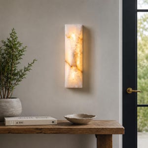 May include: A rectangular wall sconce featuring a translucent, cream-colored stone panel with natural veining. The fixture emits a warm, soft glow from within, highlighting the organic texture and patterns of the stone material against the wall.