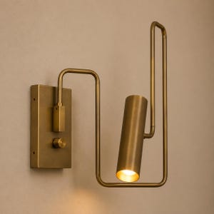 May include: A wall-mounted light fixture crafted from brushed brass metal. It features a rectangular base plate with a circular control knob and a tubular light head attached to a bent, angular frame that extends outward and upward from the wall.