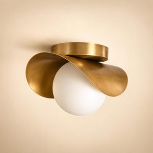May include: A flush mount ceiling light fixture featuring a spherical white glass globe nestled within a curved, brushed brass metal shade. The circular brass canopy attaches to the ceiling, supporting the wide, petal-like metallic gold reflector.