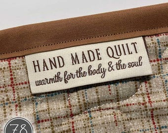 May include: A beige fabric quilt featuring a brown plaid pattern and a brown fabric border. A rectangular white label is stitched onto the quilt with the brown text "HAND MADE QUILT warmth for the body & the soul" and a circular logo with "78 makes".