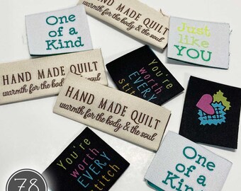 May include: A collection of rectangular fabric labels in black, white, and beige. The labels feature embroidered text including "HAND MADE QUILT warmth for the body & the soul," "One of a Kind," "Just like YOU," and "You're worth EVERY stitch."