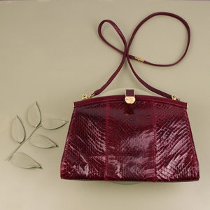 May include: A burgundy snakeskin clutch bag with a gold clasp and a long strap.