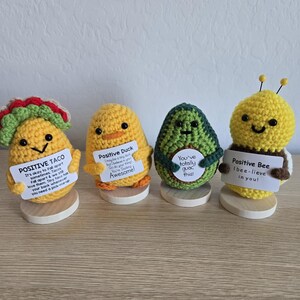 May include: Four crocheted amigurumi figures: a taco, a duck, an avocado, and a bee. Each figure holds a sign with a positive message. The taco is yellow with red and green accents. The duck is yellow with orange feet. The avocado is green. The bee is yellow and brown.