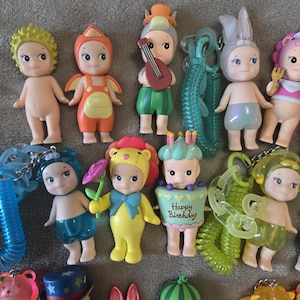 Op de afbeelding: Een verzameling kleine, kleurrijke beeldjes, elk met unieke outfits en accessoires. Sommige beeldjes zijn verkleed als dieren, terwijl andere muziekinstrumenten of bloemen vasthouden. De beeldjes zijn gemaakt van plastic en hebben een cartoonachtig uiterlijk.