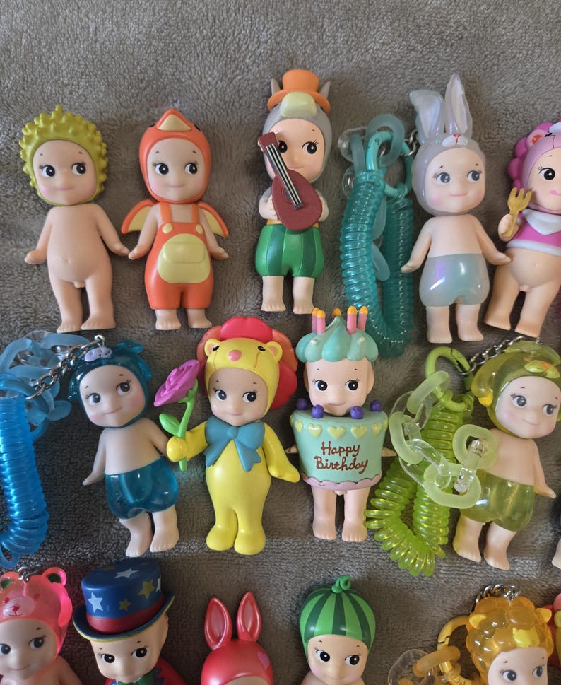 Op de afbeelding: Een verzameling kleine, kleurrijke beeldjes, elk met unieke outfits en accessoires. Sommige beeldjes zijn verkleed als dieren, terwijl andere muziekinstrumenten of bloemen vasthouden. De beeldjes zijn gemaakt van plastic en hebben een cartoonachtig uiterlijk.