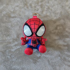 Puede incluir: Un llavero de peluche de Spider-Man con cuerpo de tela roja y azul con patr&oacute;n de telara&ntilde;a negra, grandes ojos plateados brillantes y un peque&ntilde;o emblema de ara&ntilde;a negra en el pecho. Un anillo de metal dorado est&aacute; unido a la parte superior.