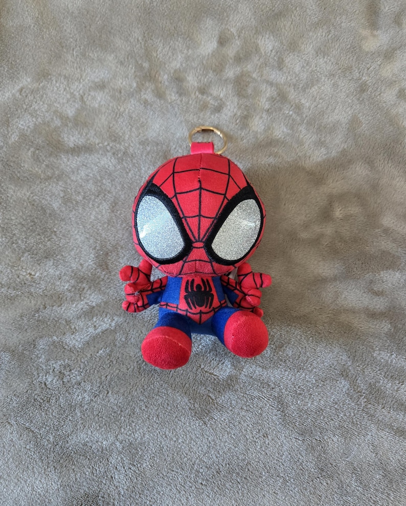 Puede incluir: Un llavero de peluche de Spider-Man con cuerpo de tela roja y azul con patr&oacute;n de telara&ntilde;a negra, grandes ojos plateados brillantes y un peque&ntilde;o emblema de ara&ntilde;a negra en el pecho. Un anillo de metal dorado est&aacute; unido a la parte superior.
