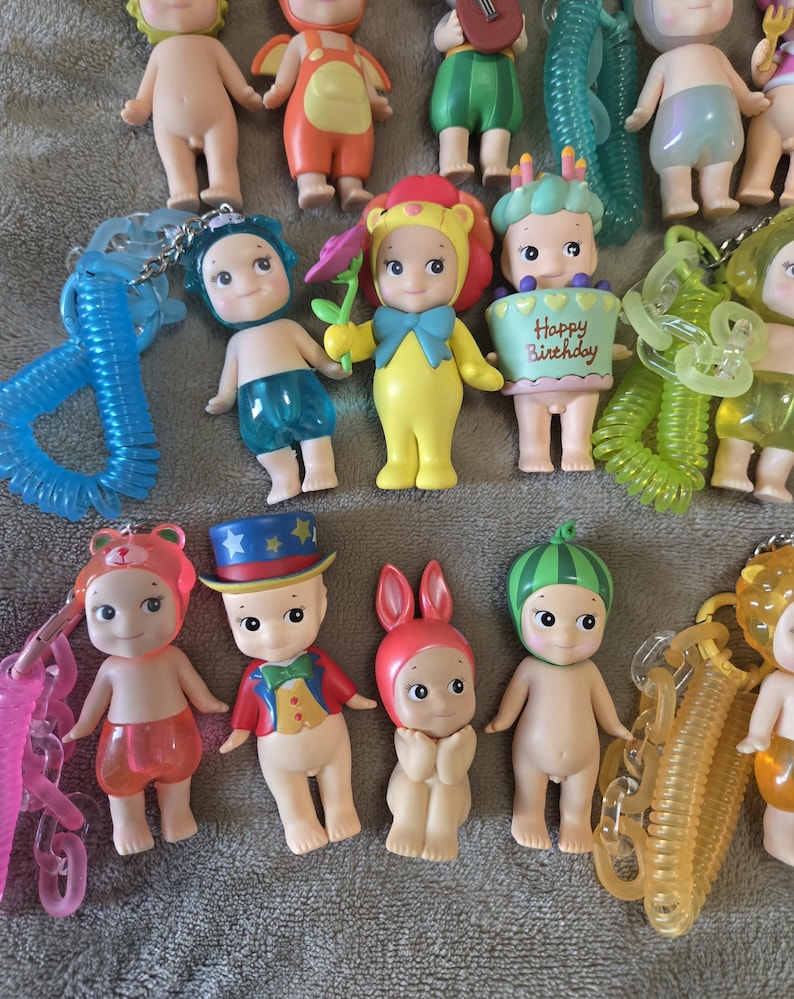 Op de afbeelding: Een verzameling kleine, decoratieve beeldjes, elk met unieke outfits en accessoires. Sommige beeldjes zijn verkleed als dieren, andere houden voorwerpen vast zoals bloemen en een taart met de tekst "Happy Birthday". De beeldjes zijn bevestigd aan sleutelhangers.