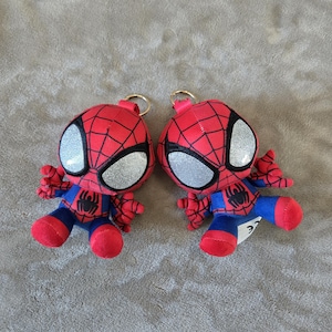 Puede incluir: Dos llaveros de peluche de Spider-Man con traje rojo y azul, patr&oacute;n de telara&ntilde;a negro, grandes ojos plateados brillantes y un peque&ntilde;o emblema de ara&ntilde;a negra en el pecho. Cada personaje tiene una anilla de metal dorado en la parte superior.