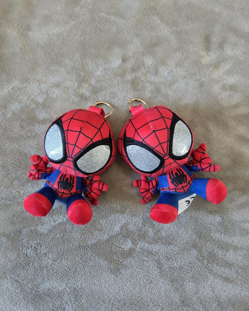 Puede incluir: Dos llaveros de peluche de Spider-Man con traje rojo y azul, patr&oacute;n de telara&ntilde;a negro, grandes ojos plateados brillantes y un peque&ntilde;o emblema de ara&ntilde;a negra en el pecho. Cada personaje tiene una anilla de metal dorado en la parte superior.