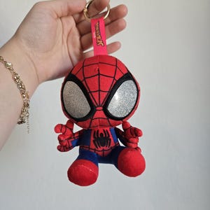 Puede incluir: Un llavero de peluche de Spider-Man que presenta un personaje rojo y azul con grandes ojos plateados brillantes, un emblema de ara&ntilde;a negro en el pecho y una correa de tela rosa con el texto Spider-Man unido a una anilla de metal dorado.