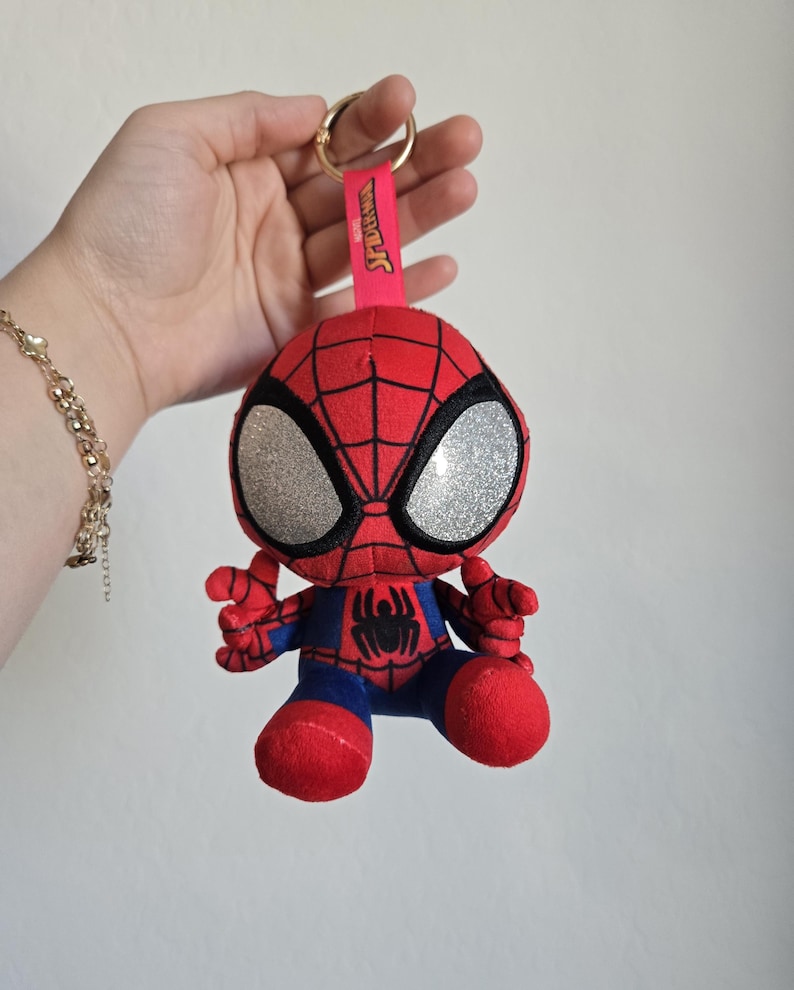 Puede incluir: Un llavero de peluche de Spider-Man que presenta un personaje rojo y azul con grandes ojos plateados brillantes, un emblema de ara&ntilde;a negro en el pecho y una correa de tela rosa con el texto Spider-Man unido a una anilla de metal dorado.