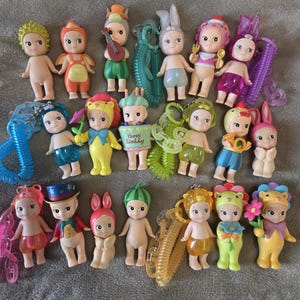 Op de afbeelding: Een verzameling kleine, grillige beeldjes met verschillende outfits en accessoires. Sommige beeldjes hebben hoeden met dierenthema's, terwijl andere muziekinstrumenten of borden vasthouden. De beeldjes zijn in verschillende kleuren, waaronder roze, geel, groen en blauw.