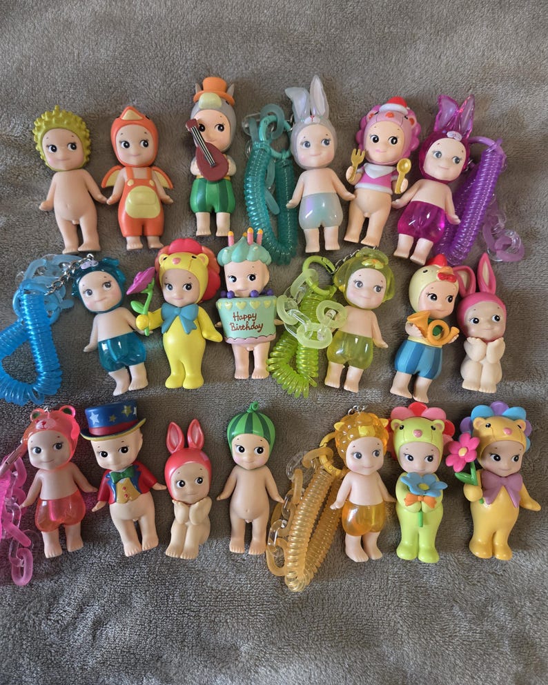 Op de afbeelding: Een verzameling kleine, grillige beeldjes met verschillende outfits en accessoires. Sommige beeldjes hebben hoeden met dierenthema's, terwijl andere muziekinstrumenten of borden vasthouden. De beeldjes zijn in verschillende kleuren, waaronder roze, geel, groen en blauw.