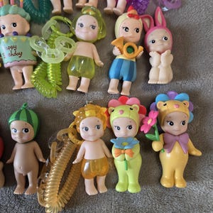 Op de afbeelding: Een verzameling kleine, grillige beeldjes met verschillende outfits en accessoires. Sommige beeldjes zijn verkleed als dieren, terwijl andere muziekinstrumenten of bloemen vasthouden. Het kleurenpalet omvat pasteltinten en heldere tinten, met een focus op schattige en speelse ontwerpen.
