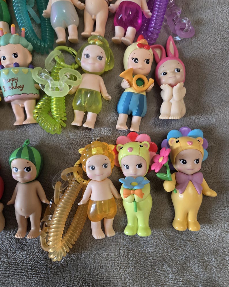Op de afbeelding: Een verzameling kleine, grillige beeldjes met verschillende outfits en accessoires. Sommige beeldjes zijn verkleed als dieren, terwijl andere muziekinstrumenten of bloemen vasthouden. Het kleurenpalet omvat pasteltinten en heldere tinten, met een focus op schattige en speelse ontwerpen.