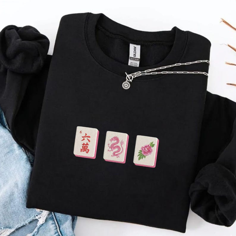 May include: Black sweatshirt featuring three embroidered Mahjong tiles with pink accents. The tiles display Chinese characters, a dragon, and a flower. A silver chain necklace with a pendant is draped across the sweatshirt.