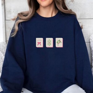 May include: Navy blue sweatshirt with three embroidered Mahjong tiles. The tiles feature a red bow, bamboo stalks, and a floral design. The word "Joker" is above the bow tile.