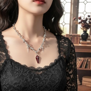 May include: A silver necklace with a barbed wire design and small dagger pendants. The necklace has a coffin-shaped pendant with a red centre and a cross. The necklace is worn over a black lace top.