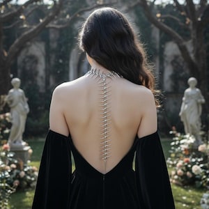 May include: A silver spine-shaped necklace is worn on the back of a person in a black velvet dress with long sleeves. The necklace runs from the neck down the spine. The background shows a garden with statues and trees.