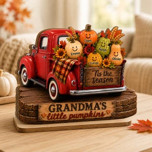 Puede incluir: Una figura de camión rojo de estilo vintage que transporta cinco calabazas sonrientes con los nombres Emma, Liam, Noah, Lucas y Olivia. La caja del camión tiene una manta a cuadros y el texto 'Tis the season' sobre una base 'GRANDMA'S little pumpkins'.
