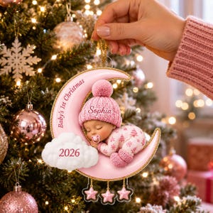 K&ouml;nnte beinhalten: Ein rosafarbener Weihnachtsbaumschmuck in Form einer Mondsichel mit einem schlafenden Baby in einer rosa Strickm&uuml;tze und einem gemusterten Strampler. Der Mond tr&auml;gt die Aufschrift Baby's 1st Christmas und Sophia sowie eine Wolke mit dem Jahr 2026.