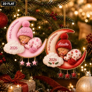 K&ouml;nnte beinhalten: Zwei halbmondf&ouml;rmige Anh&auml;nger mit schlafenden Babys in Strickm&uuml;tzen und Schlafanz&uuml;gen. Einer ist rosa mit dem Namen Sophia, der andere rot mit Amelia. Beide tragen die Aufschrift Baby's 1st Christmas, das Jahr 2026 und h&auml;ngende Sterne.