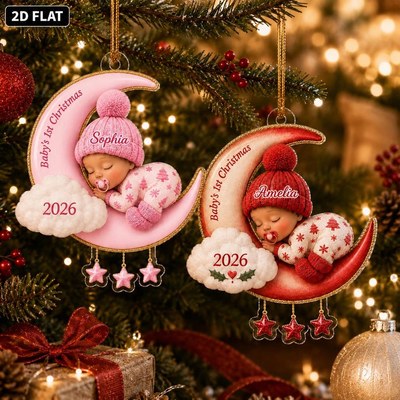 K&ouml;nnte beinhalten: Zwei halbmondf&ouml;rmige Anh&auml;nger mit schlafenden Babys in Strickm&uuml;tzen und Schlafanz&uuml;gen. Einer ist rosa mit dem Namen Sophia, der andere rot mit Amelia. Beide tragen die Aufschrift Baby's 1st Christmas, das Jahr 2026 und h&auml;ngende Sterne.