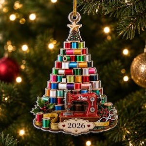 Puede incluir: Un adorno en forma de árbol de Navidad con carretes de hilo coloridos apilados, una máquina de coser roja con el nombre Mimi, una cinta métrica amarilla y una placa en la base con el año 2026, rematado con una estrella dorada.