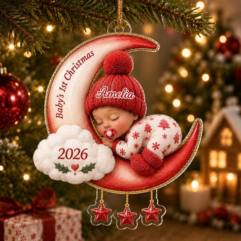 K&ouml;nnte beinhalten: Ein Weihnachtsschmuck mit einem schlafenden Baby in roter Strickm&uuml;tze und wei&szlig;em Schneeflocken-Schlafanzug, das auf einer roten Mondsichel liegt. Der Mond zeigt den Text Baby's 1st Christmas, eine Wolke mit 2026 und den Namen Amelia auf der M&uuml;tze.