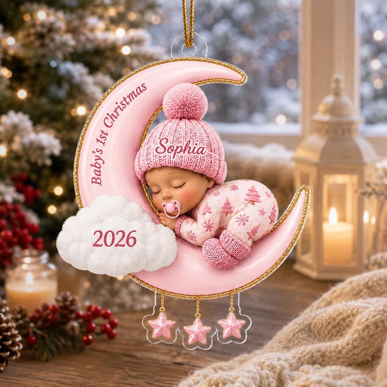 K&ouml;nnte beinhalten: Ein rosafarbener Mondsichel-Anh&auml;nger mit einem schlafenden Baby in einer rosa Strickm&uuml;tze und einem Strampler mit Schnuller. Der Mond ist mit Baby's 1st Christmas und Sophia beschriftet, mit 2026 auf einer wei&szlig;en Wolke. Drei kleine Sterne h&auml;ngen unten.