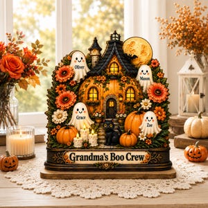 Könnte beinhalten: Eine dekorative Halloween-Tischdekoration mit einem kürbisförmigen Haus, einer schwarzen Katze und vier Geisterfiguren mit den Namen Oliver, Mason, Grace und Zoe. Der Sockel trägt die Aufschrift Grandma's Boo Crew, umgeben von orangefarbenen Blumen und Kürbissen.