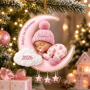 K&ouml;nnte beinhalten: Ein rosafarbener Mondsichel-Anh&auml;nger mit einem schlafenden Baby in einer rosa Strickm&uuml;tze und einem Schlafanzug mit Baummuster. Der Mond tr&auml;gt die Aufschrift Baby's 1st Christmas und Sophia, eine wei&szlig;e Wolke zeigt das Jahr 2026.