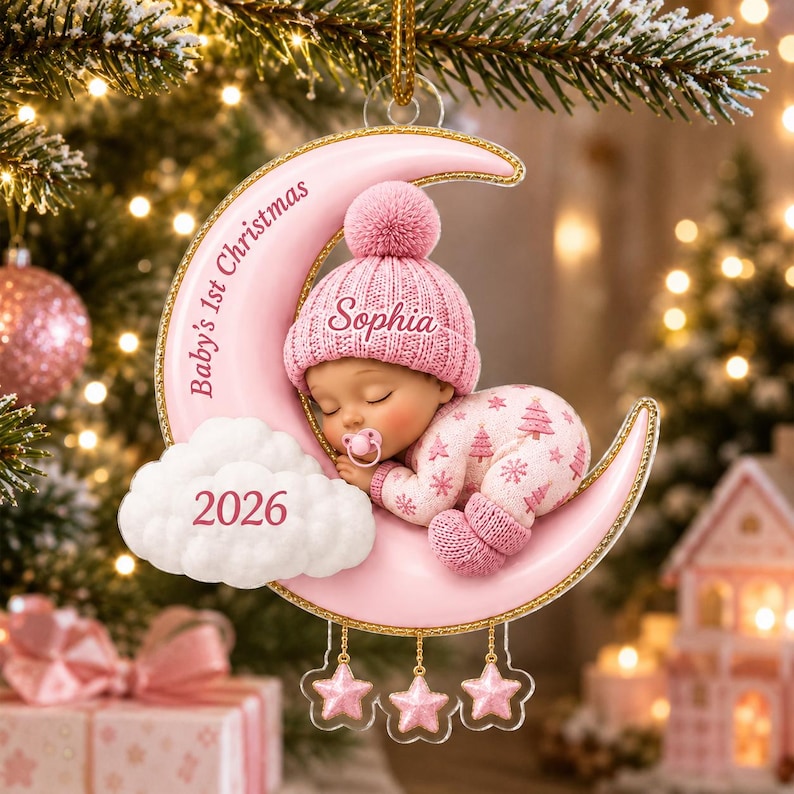 K&ouml;nnte beinhalten: Ein rosafarbener Mondsichel-Anh&auml;nger mit einem schlafenden Baby in einer rosa Strickm&uuml;tze und einem Schlafanzug mit Baummuster. Der Mond tr&auml;gt die Aufschrift Baby's 1st Christmas und Sophia, eine wei&szlig;e Wolke zeigt das Jahr 2026.