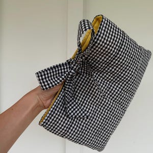 May include: A rectangular fabric pouch featuring a black and white gingham check pattern on the exterior. The pouch includes a matching fabric tie closure on the side and a solid yellow fabric lining visible along the top opening edge.
