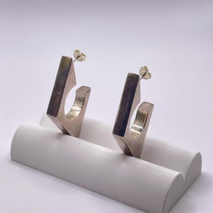May include: Two silver-coloured, geometric earrings with a square design and post backs. The earrings are displayed on a white, curved jewellery holder. The earrings have a modern, minimalist aesthetic.