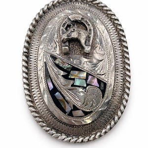 May include: An oval-shaped silver-toned belt buckle featuring a rope-style border, an engraved horse head inside a horseshoe, and a decorative inlay of iridescent abalone shell pieces set against a dark background with intricate metal scrollwork.