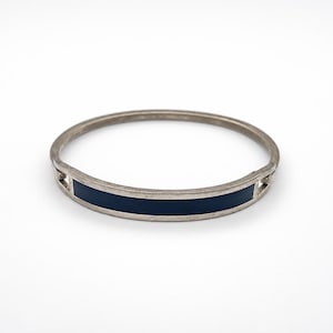 Peut inclure: Un bracelet jonc en métal argenté doté d'une bande incrustée bleu marine foncé lisse centrée sur la partie avant. Le bracelet présente une finition métallique polie avec de petites découpes rectangulaires ouvertes de chaque côté du panneau bleu.