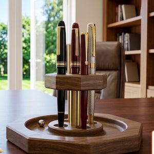 May include: An octagonal wooden pen stand with a central pillar and slots holding four fountain pens in black, burgundy, and silver finishes. A separate burgundy fountain pen with a gold nib lies on the surface next to the stand.