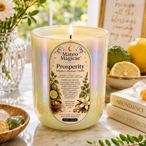 Puede incluir: Una vela en frasco de vidrio iridiscente con una mecha encendida, con una etiqueta que dice Mateo Magicae Prosperity, Bergamot, Patchouli, Vanilla, 8 OZ | 226 G. La etiqueta incluye ilustraciones botánicas de cítricos y flores de vainilla.