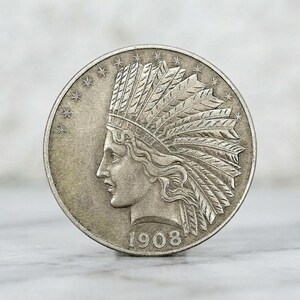 May include: A silver-toned circular coin featuring a profile of a person wearing a feathered headdress. The word LIBERT is inscribed on the headband, and the year 1908 is printed at the bottom. Thirteen stars are arranged along the upper left edge.
