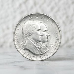 Puede incluir: Una moneda conmemorativa de los Estados Unidos de color plateado con los retratos de perfil de George Washington y otra figura. El texto LIBERTY, UNITED STATES OF AMERICA e IN GOD WE TRUST está grabado, junto con una marca de ceca S.